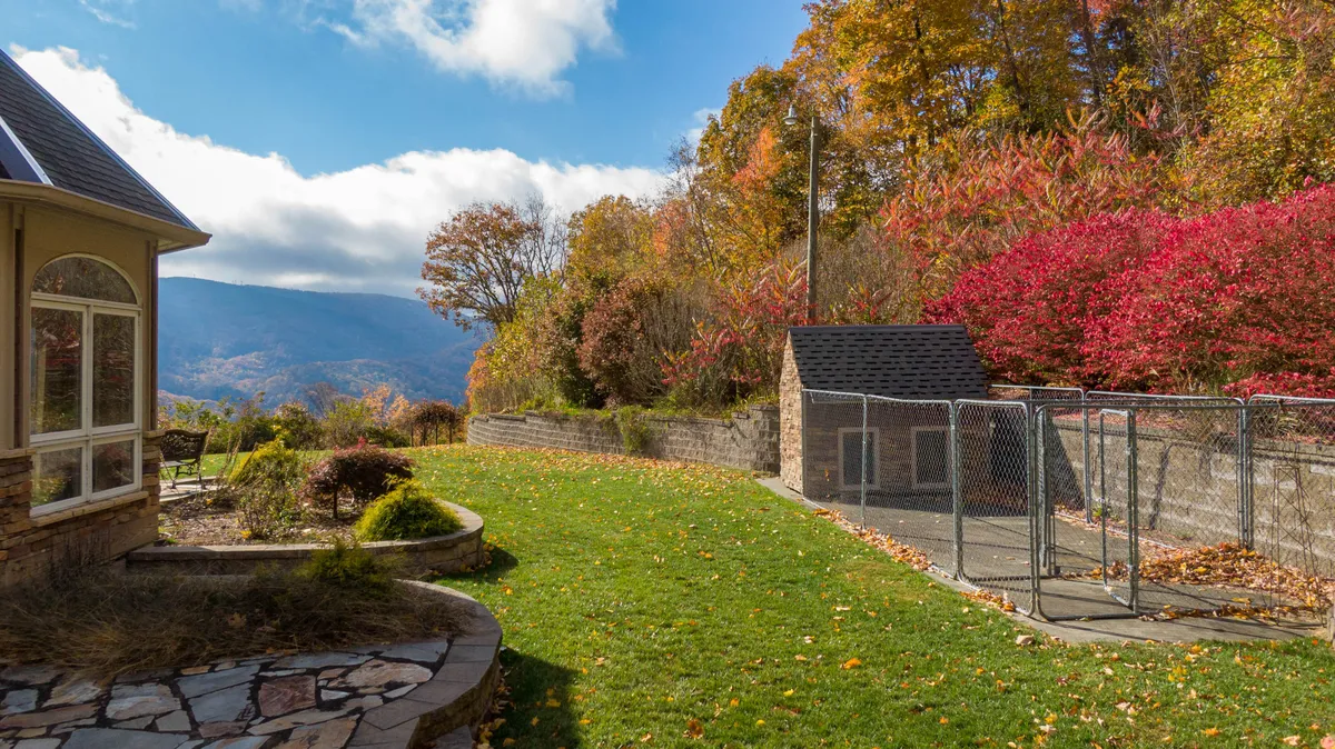 A stone pathway winds through a lush green lawn and vibrant autumn foliage, leading to a fenced area that could serve as a secure play spot for pets. The landscape shows the expansive outdoor area and the mountain view in the distance.