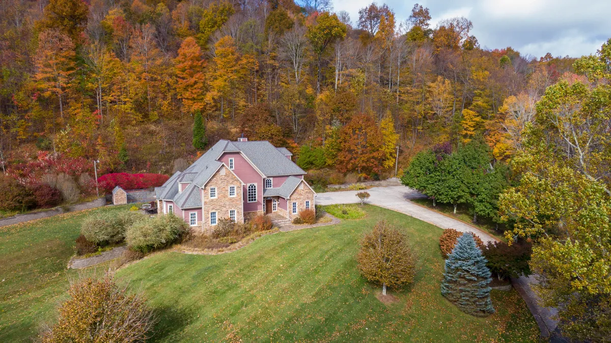 A serene exterior view of Mountain Gem, featuring the multi-level house nestled among mature trees with a spacious driveway leading up to the property. The porch offers glimpses of outdoor gathering spaces, surrounded by vibrant greenery—perfect for family activities and enjoying the natural setting.
