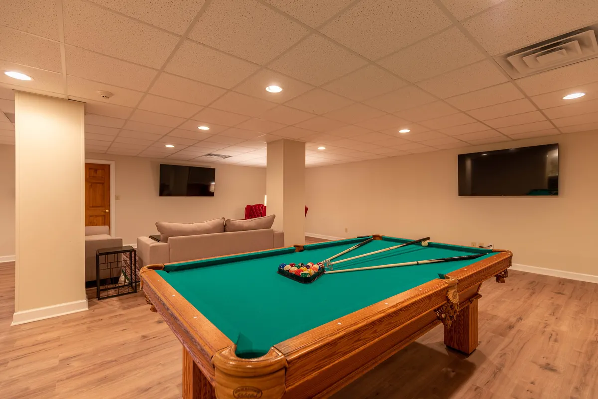 A vibrant game room centers on a sleek pool table, with a cozy sofa and large wall-mounted TV nearby. The setting is modern yet inviting, perfect for gathering with friends or family for spirited games, movie nights, or cheering on a favorite sports team. Soft lighting and warm color tones enhance the welcoming, playful atmosphere.