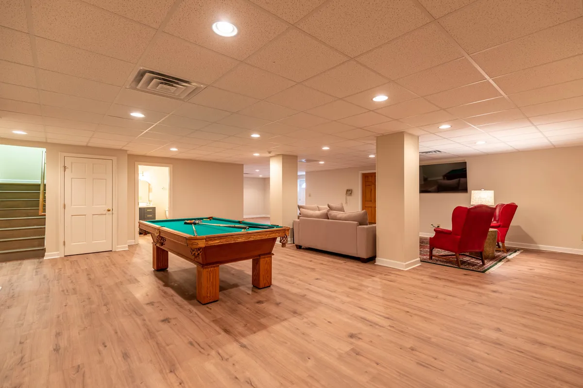 A dynamic angle of the game room highlights a pool table at the center, flanked by vibrant red armchairs, a plush sofa, and a large TV mounted on the wall. The open layout encourages both active play and relaxed spectating, while the bold colors inject energy into the space.