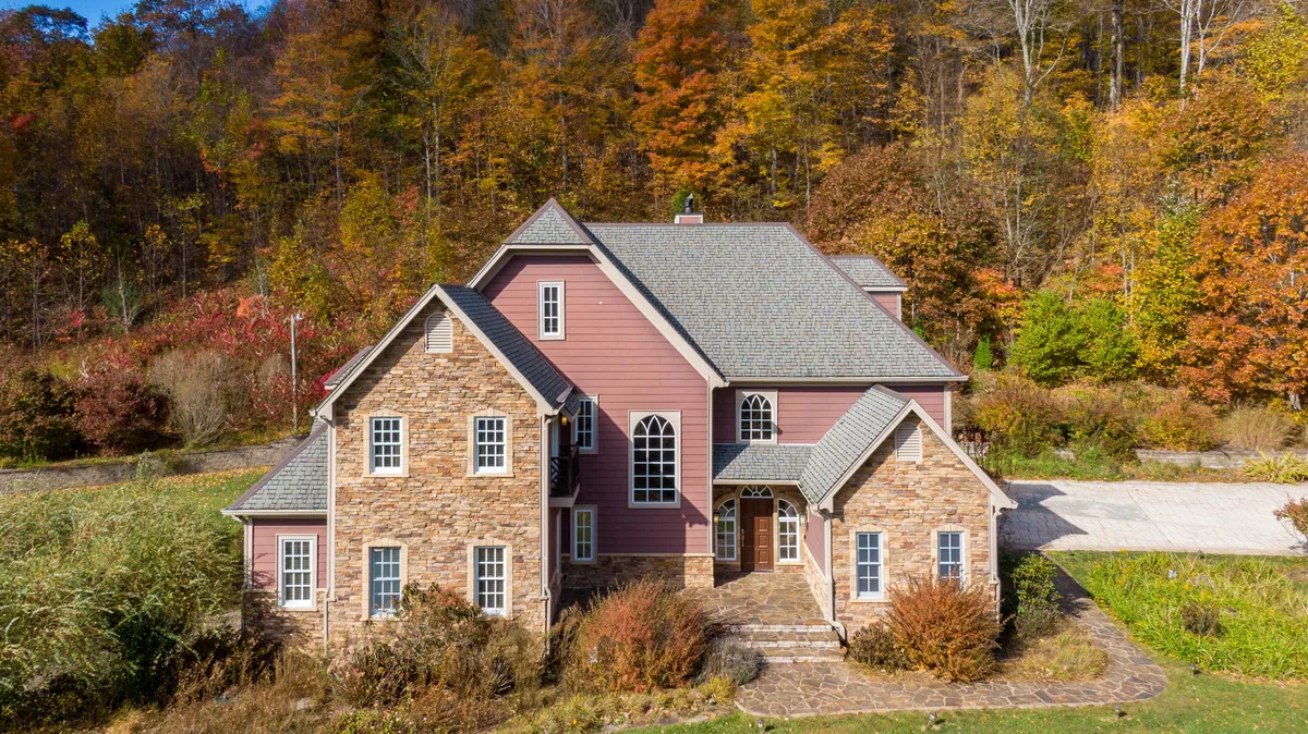 The exterior of Mountain Gem features a striking stone facade and gabled roof nestled among lush trees and foliage. Large windows offer glimpses of the inviting interior, while the surrounding landscape transitions beautifully through all four seasons.