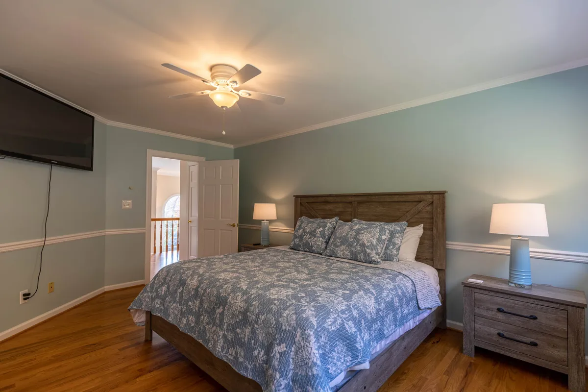A serene king bedroom featuring a comfortable bed with patterned bedding, a nightstand with a lamp, and a wall-mounted TV. Natural light streams through the windows, creating a restful and inviting atmosphere.