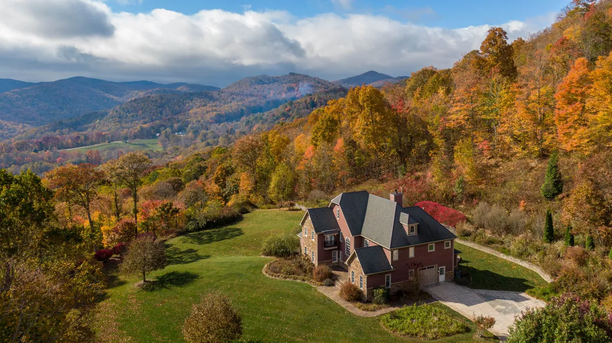 The image showcases a grand, multi-story home perched atop a mountain, surrounded by vibrant, colorful trees with an expansive view of the Blue Ridge Mountains in the distance. The setting sun casts a golden glow over the landscape, evoking a tranquil and inviting atmosphere perfect for gatherings and relaxation.
