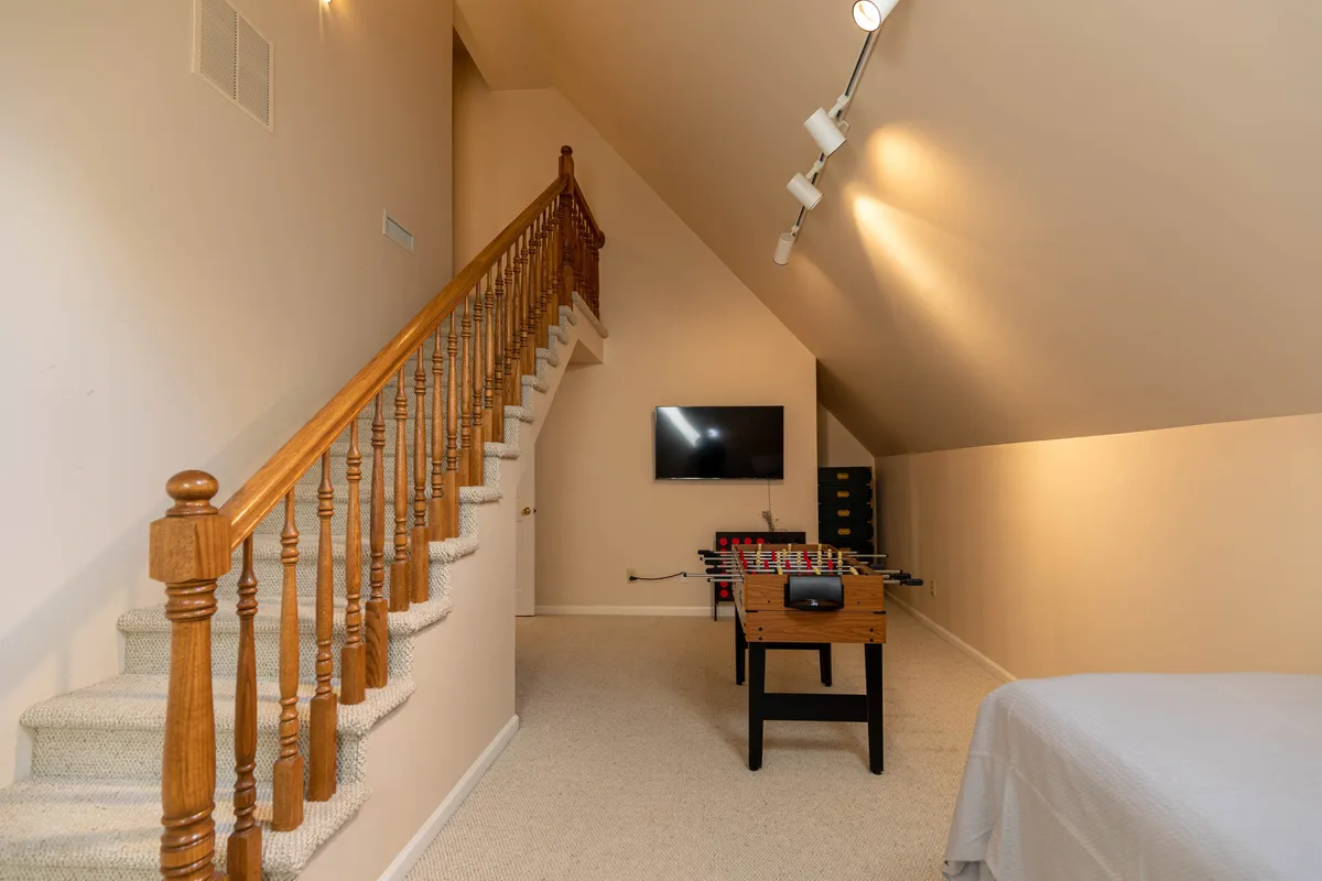 A playful lower level bedroom features a foosball table at the foot of a cozy bed, with a large wall-mounted TV and a staircase leading to other parts of the home. The room blends restful comfort with energetic fun, making it an ideal spot for kids or adults to unwind and compete.