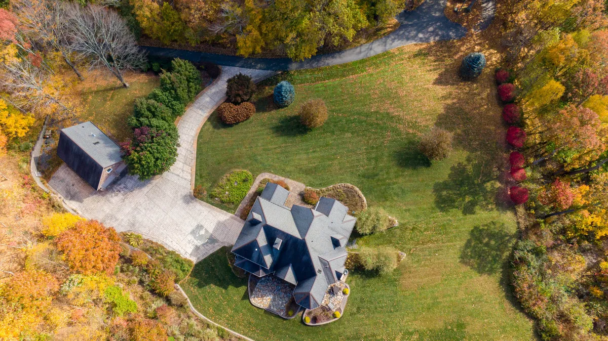 The main house is visible beyond a sweeping driveway, nestled among lush trees and featuring an adjacent outbuilding. The well-maintained garden and outdoor space invite a variety of activities, from family gatherings to casual exploration with pets.