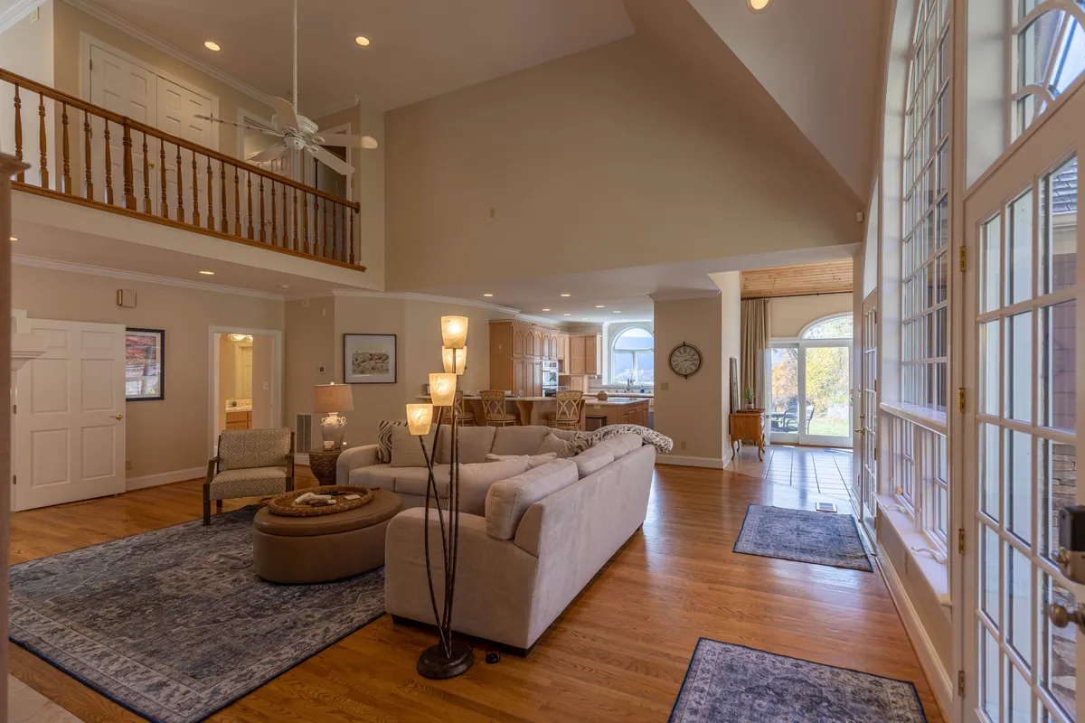 This image captures a spacious open-plan living area featuring a large sectional sofa, tall windows that bathe the space in sunlight, a visible dining area, and a modern kitchen with decorative lighting. The layout highlights the seamless flow between lounging, dining, and cooking spaces, set under high ceilings.