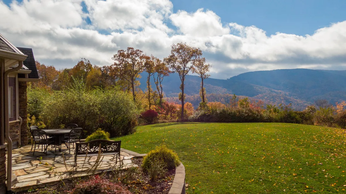 A stone patio furnished with a table and chairs offers an ideal alfresco dining space, overlooking distant mountains. Colorful foliage frames the scene, suggesting year-round beauty and tranquil outdoor living.