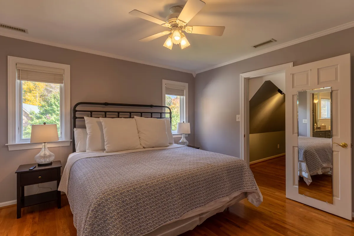 A bright, elegant bedroom with a stylish bed adorned with decorative pillows, nightstands with lamps on either side, large windows filling the room with sunlight, and a mirror on the closet door. The mood is serene and comfortable, ideal for unwinding after a day outside.