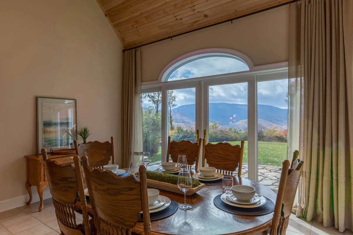 A dining area with a rustic wooden table set for a meal, surrounded by wooden chairs and framed by large windows with mountain views. The space is filled with natural light, creating a warm, festive mood perfect for shared celebrations.